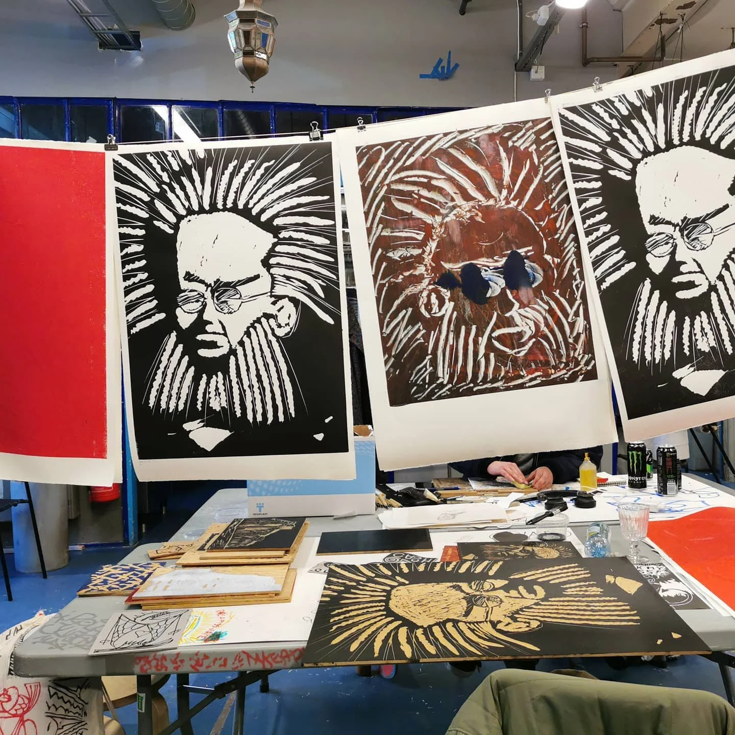 To personer som lager kunstverk av store, fargede portretter av en mann med solhatt og briller, i et kunstverksted med arbeidsbord, utstyr og malerier.