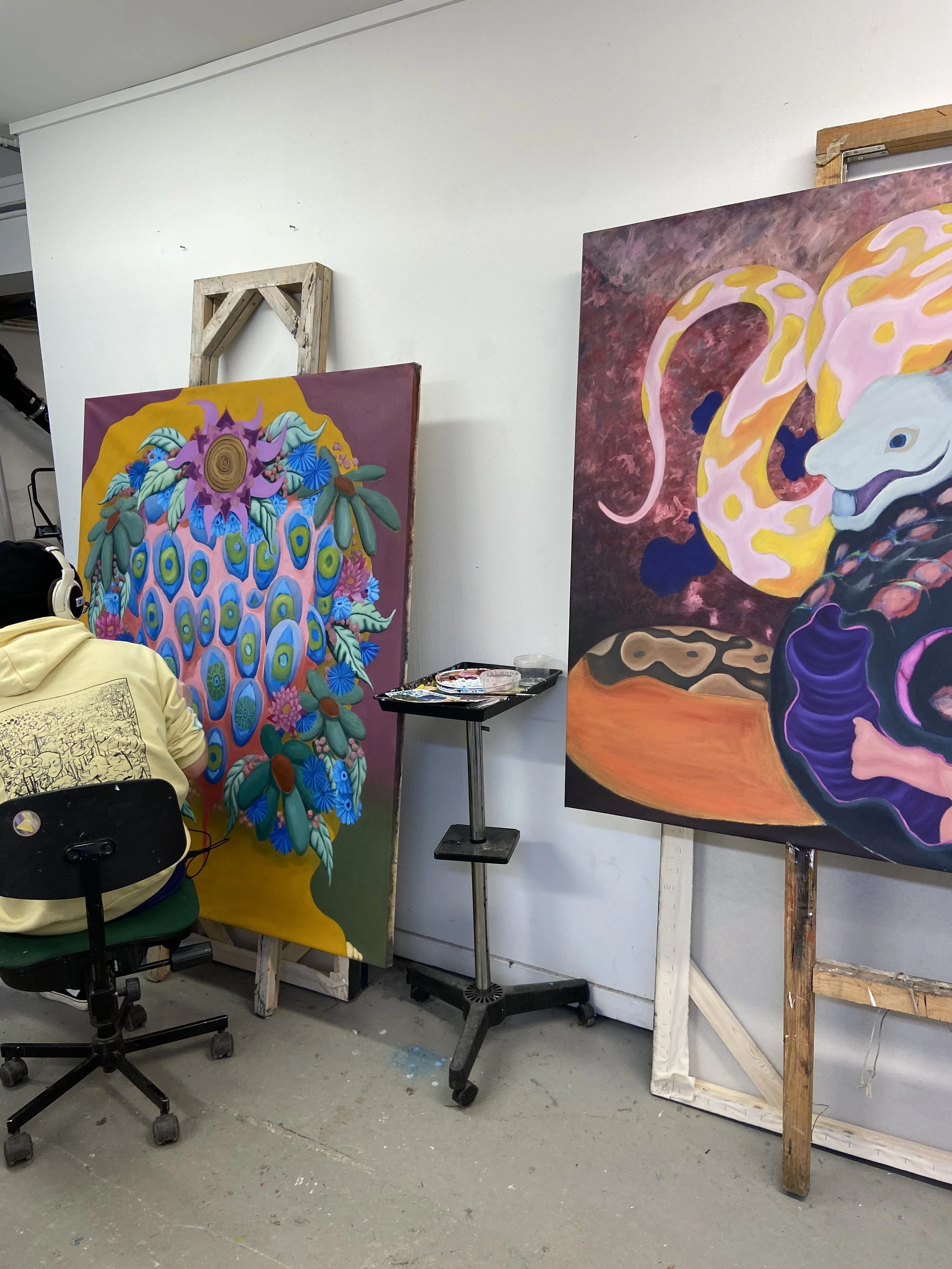 To malerier på lerret i et studio, en maleri viser en fargerik figur med palmeblader og øyne, det andre viser en hvit drage med gul og rosa mønster, omgitt av mørkere bakgrunnsdetaljer.