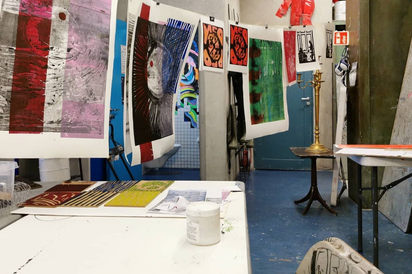 En kunststudio med utskrifter av ulike kunstverk hengende på en snor. Det er maler, stiger og et bord med kunstmaterialer.