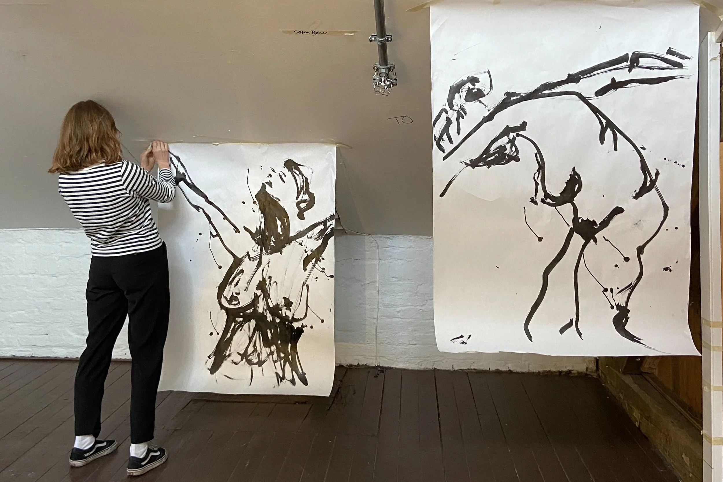 En person med stripete genser som maler store svarte abstrakte linjetegninger på store ark med hvitt papir.