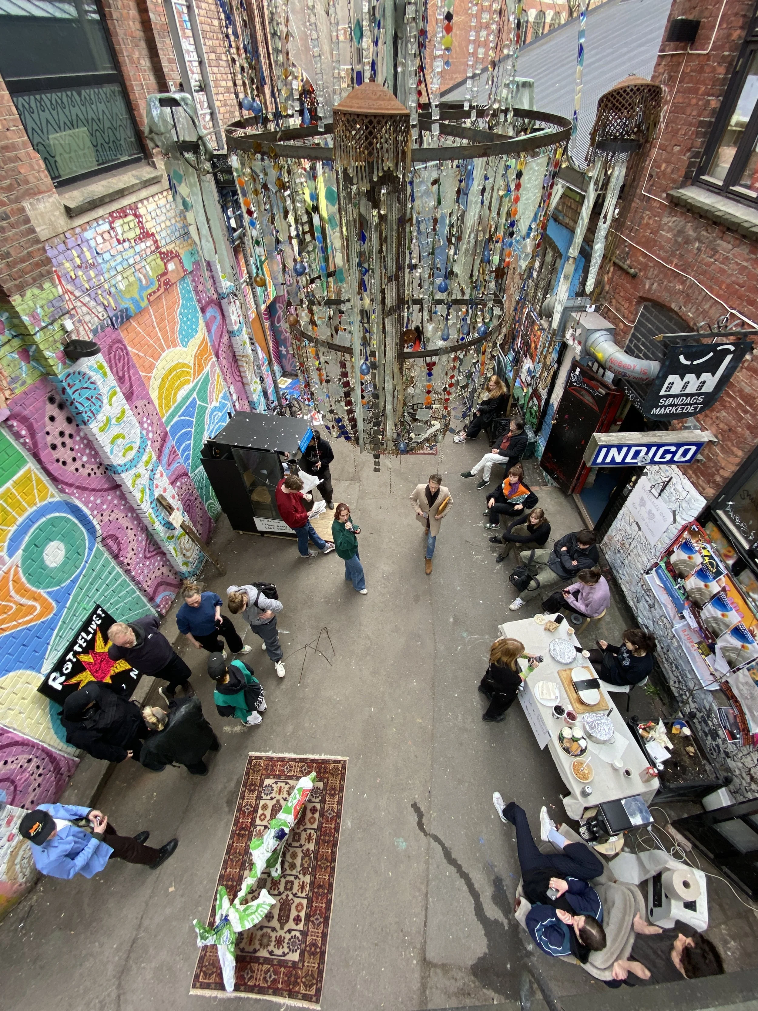 Utsikt over en liten gårdsplass med mennesker som sosialiserer, det er malt fargerike vegger og et stort kunstinstallasjon av hengende fargede glass eller plastobjekter i midten.