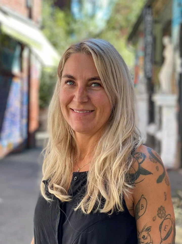 En blond kvinne som smiler, kledd i svart topp, med tatoveringer på venstre arm, står utendørs med bakgrunn av fargerike veggmalerier og trær.