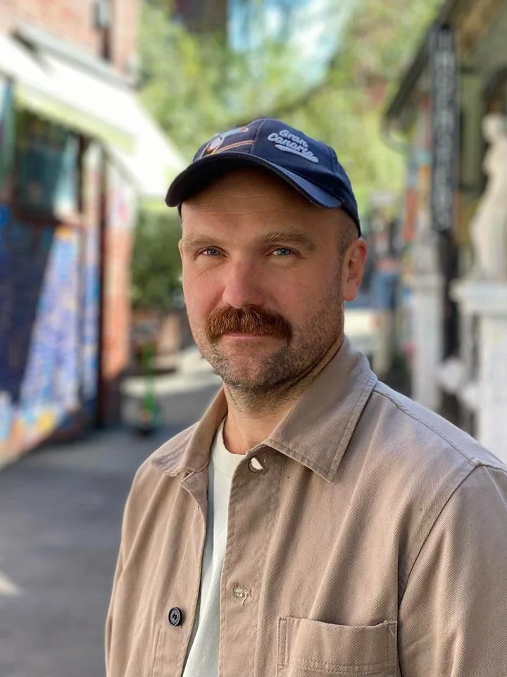 Man med skjegg og baseballhatt står på en gate i en by, bakgrunn med fargerike murvegger og noen trær.