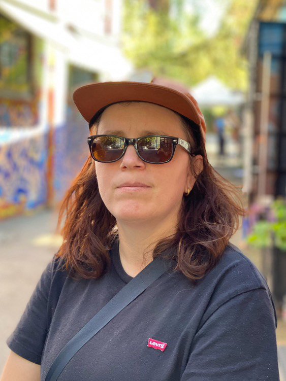 Kvinne med brunt basest, mørke solbriller og svart t-skjorte som står utendørs med fargerik vegg i bakgrunnen.