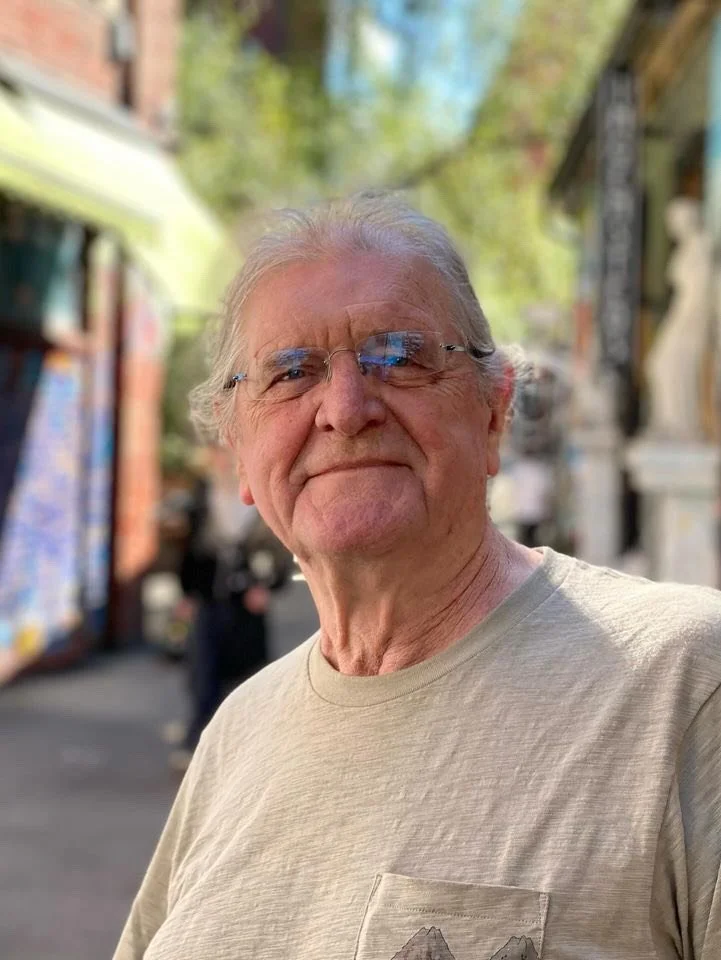 En eldre mann med grått hår, iført briller og en beige T-skjorte står utenfor på en gate i solskinn, med butikker og gjenstander i bakgrunnen.