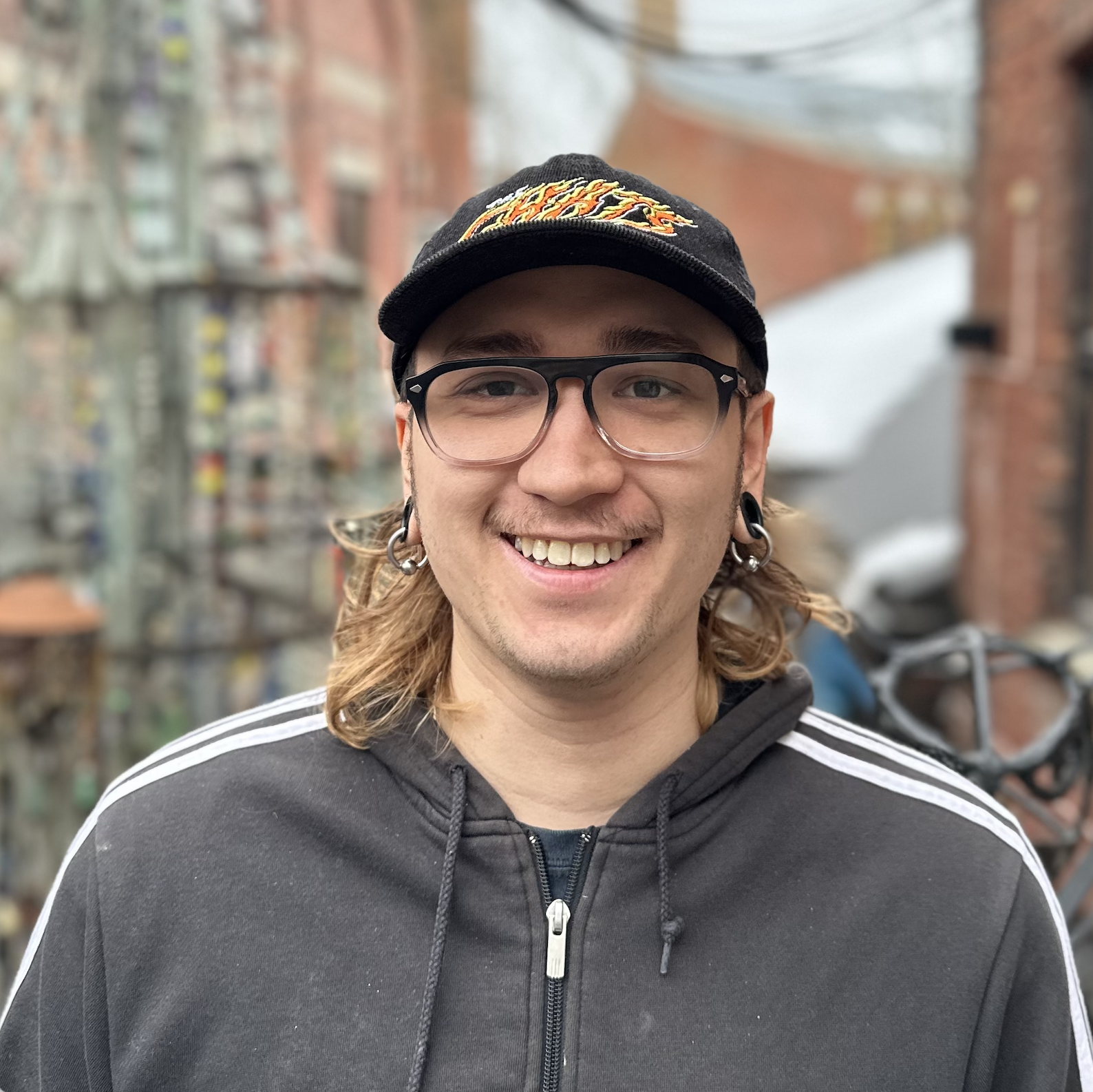 En ung mann med langt hår, iført briller, øredobber og caps, smiler foran en bygate. Han har på seg en svart hoodie med hvite striper.
