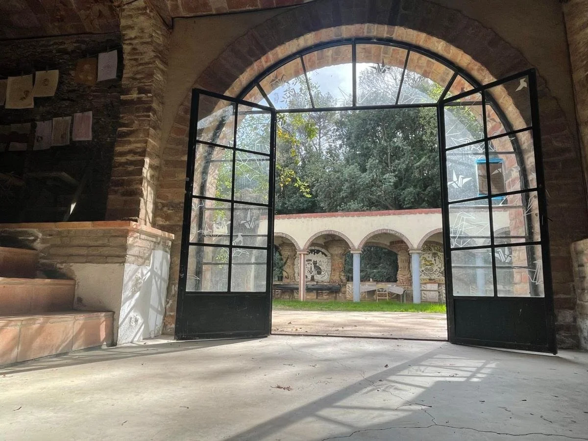 Utsikt gjennom åpne sorte dører mot en åpen gårdsplass med arkader og graffiti på veggene, omgitt av trær.