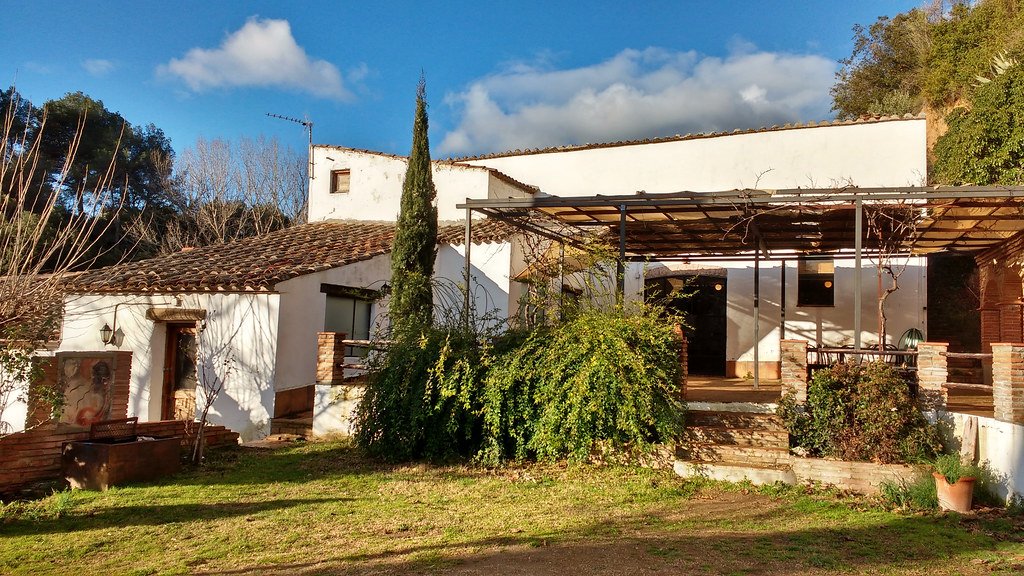 Hus med hvite vegger, terrasser, trær, planter og en blå himmel med noen skyer.