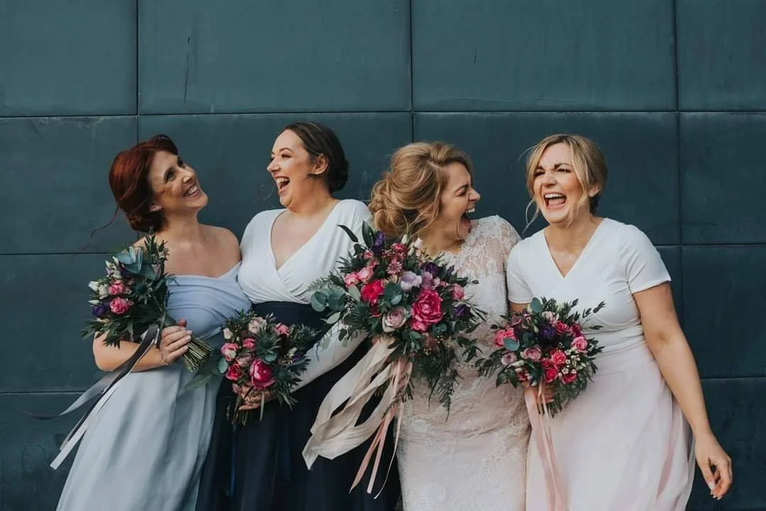 Four women in wedding and bridesmaid dresses holding bouquets, laughing together outdoors.