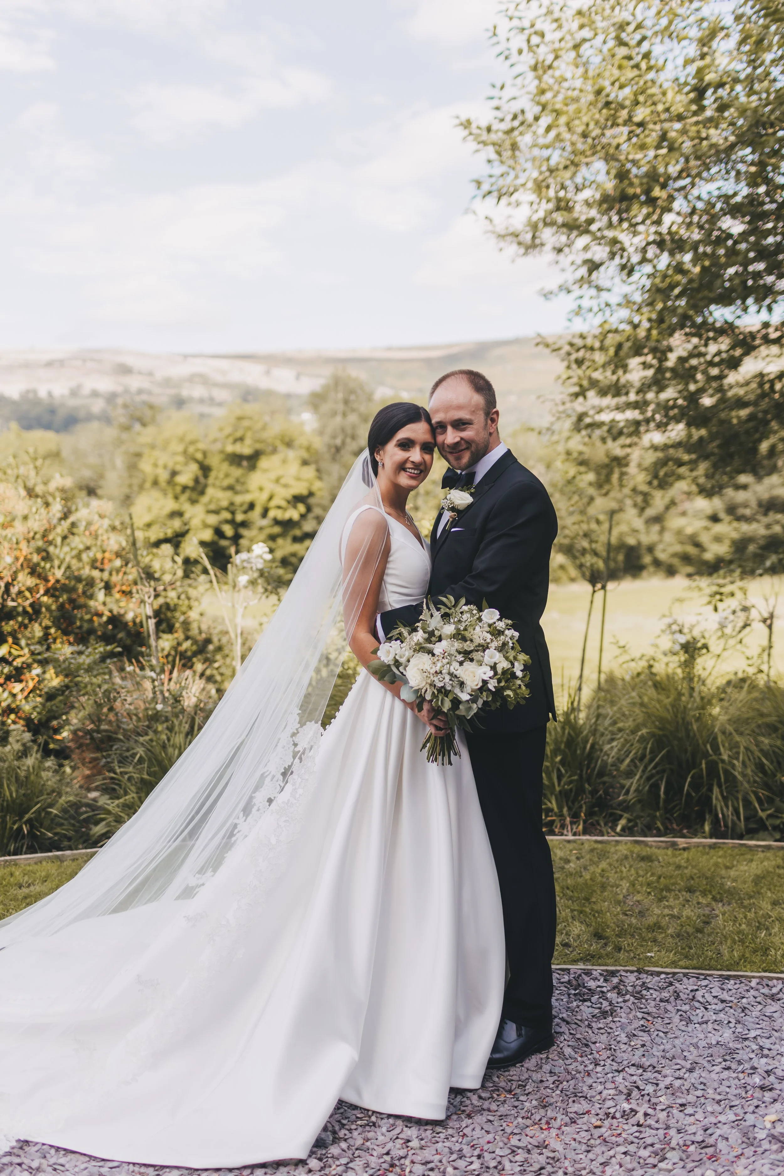 A bride and groom smiling together outdoors on their wedding day, with the bride holding a bouquet of white flowers and the groom dressed in a tuxedo, against a scenic green landscape with trees and a cloudy sky.