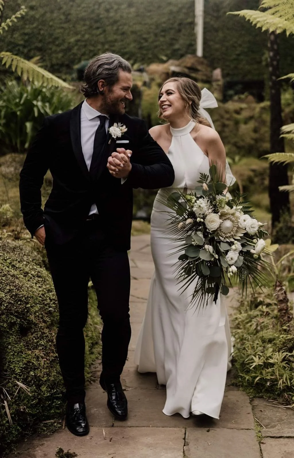 A bride and groom walking outdoors, smiling and holding hands, surrounded by lush greenery, with the bride carrying a large bouquet of white flowers and greenery.