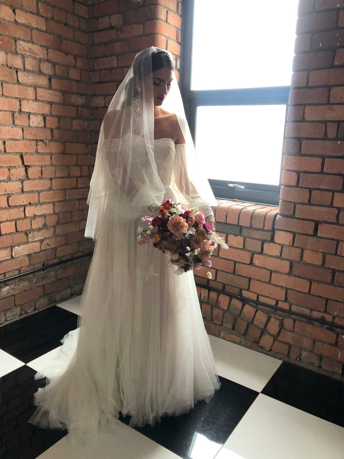 Bride dressed in a white wedding gown with an off-the-shoulder design and a long tulle veil, holding a colorful bouquet of flowers, standing near a large window with brick walls in the background.
