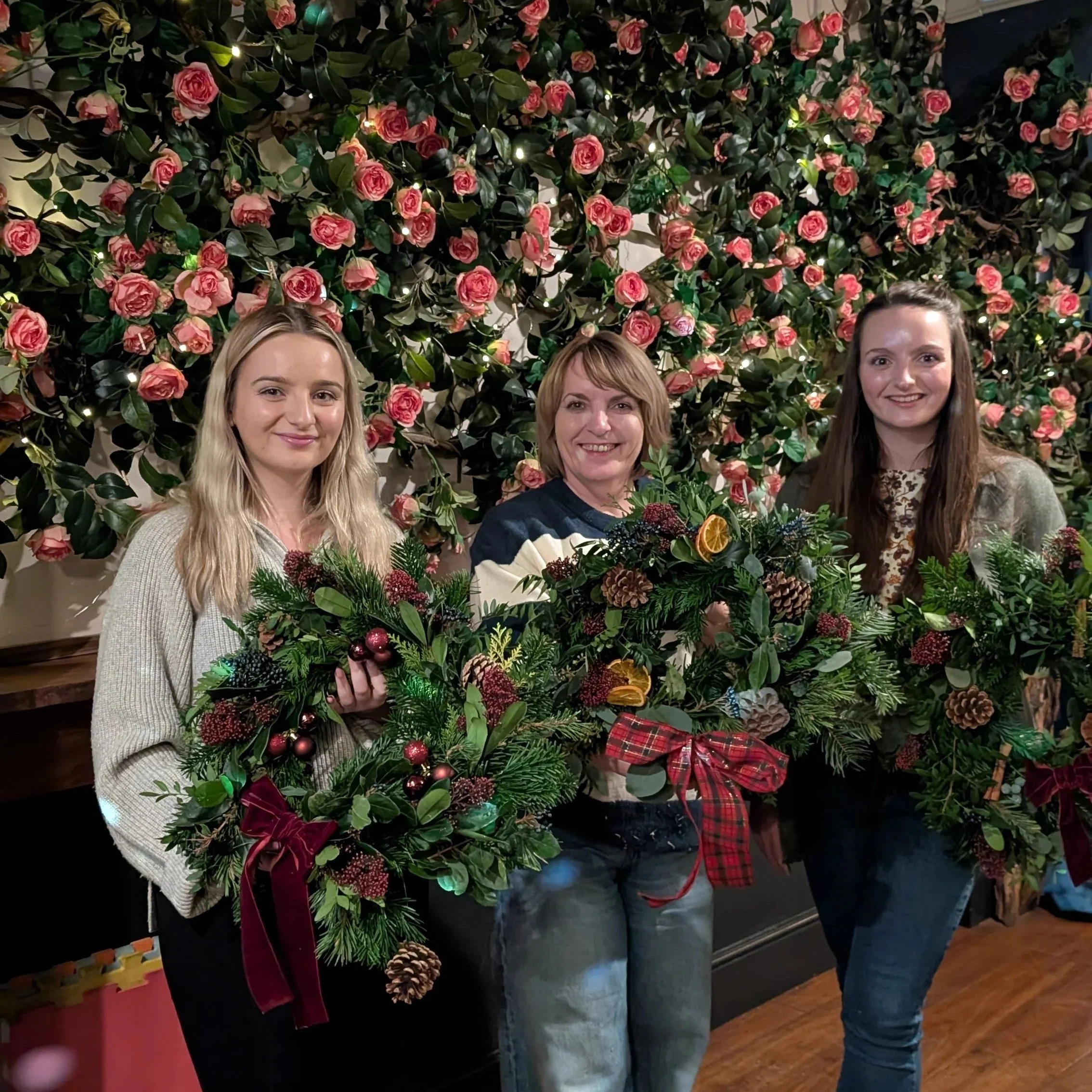 Three women standing in front of a wall decorated with pink roses, holding Christmas wreaths with pine, berries, pinecones, dried orange slices, and ribbons.