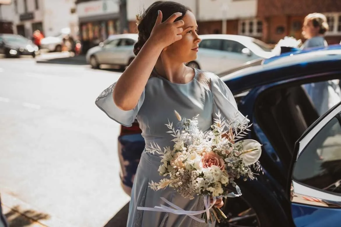 A woman in a light gray dress holding a bouquet of flowers, standing next to a car, on a city street during daytime.