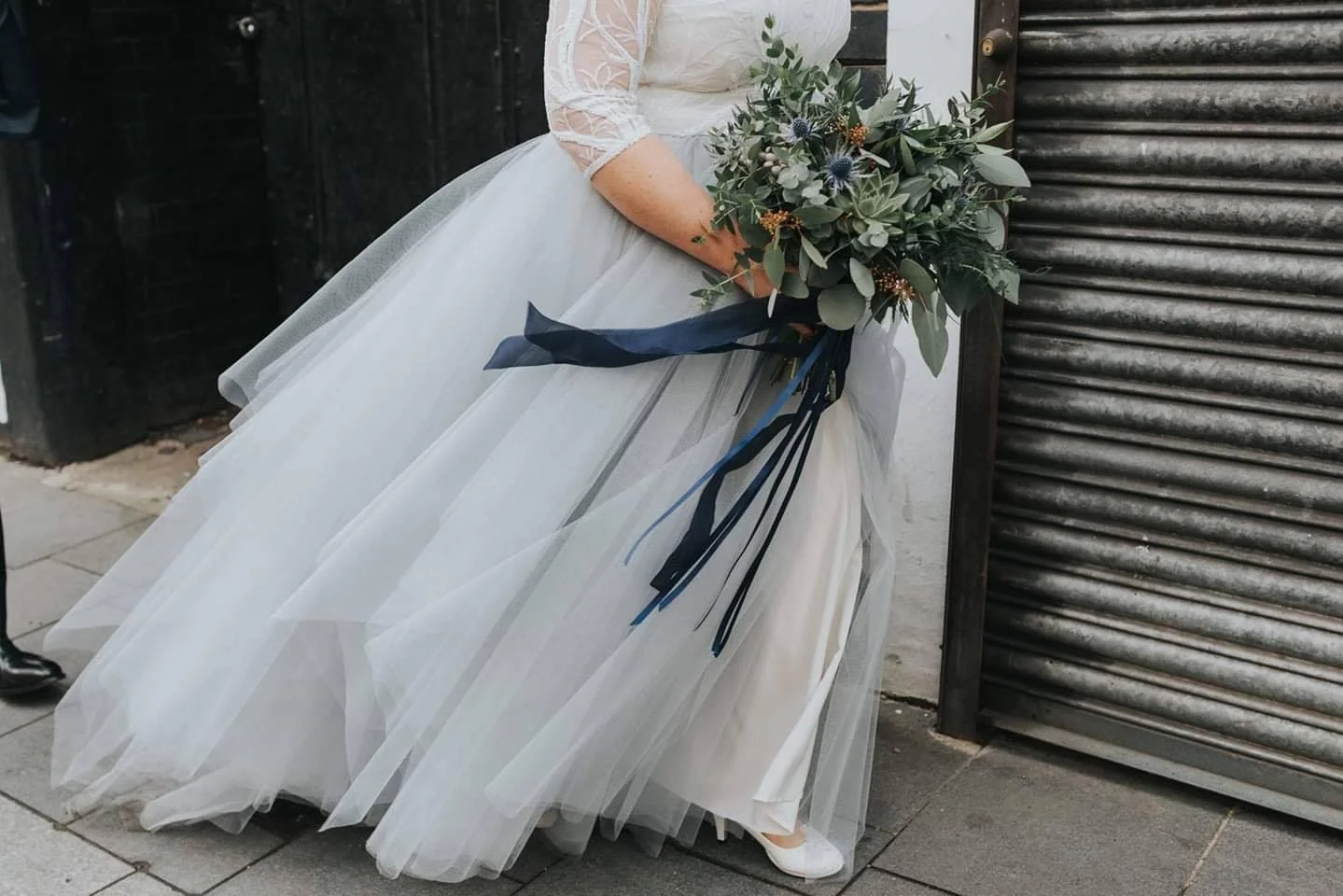 A bride in a white wedding dress holding a bouquet of greenery and flowers, standing outdoors next to a black metal gate.