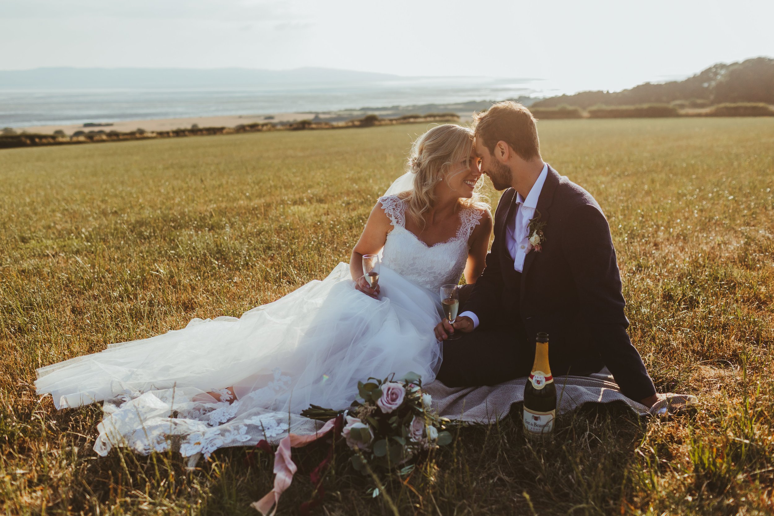 A bride and groom sitting in a grassy field during sunset, smiling and leaning their foreheads together, holding champagne glasses, with a bouquet and a bottle of champagne nearby.