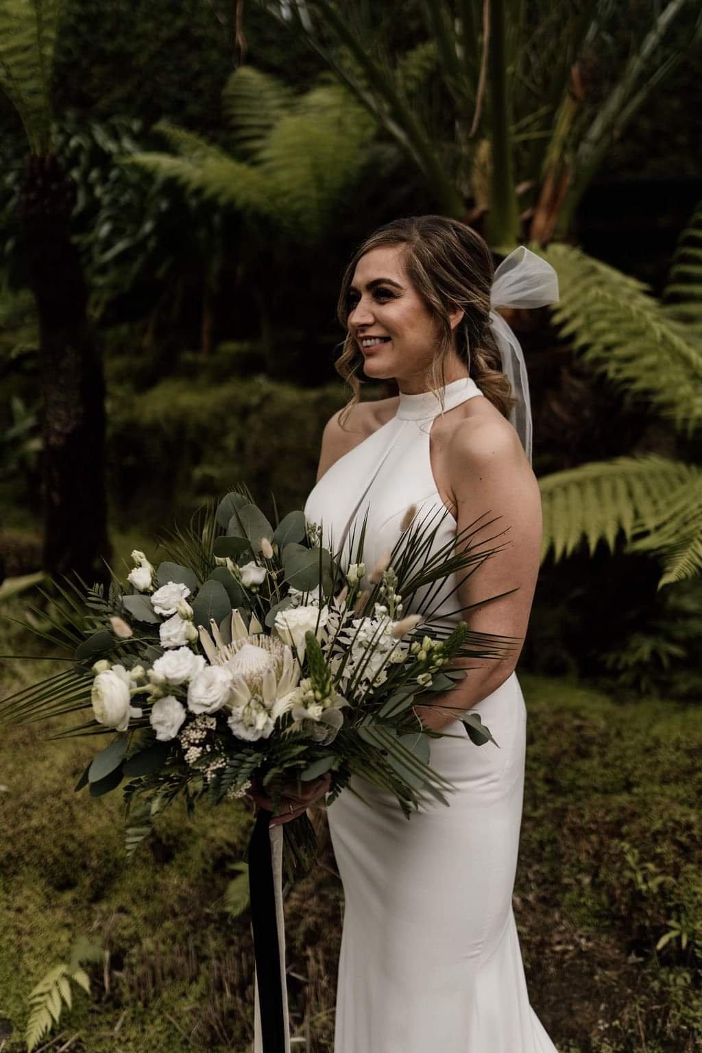 A smiling woman in a white dress with a ribbon in her hair holding a large bouquet of white flowers and greenery, standing in a lush, green outdoor setting with fern plants in the background.