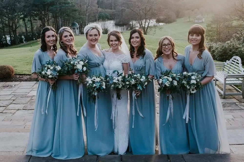 A bride and six bridesmaids standing outdoors on a stone path, all dressed in matching light blue dresses and holding bouquets of white and blue flowers.