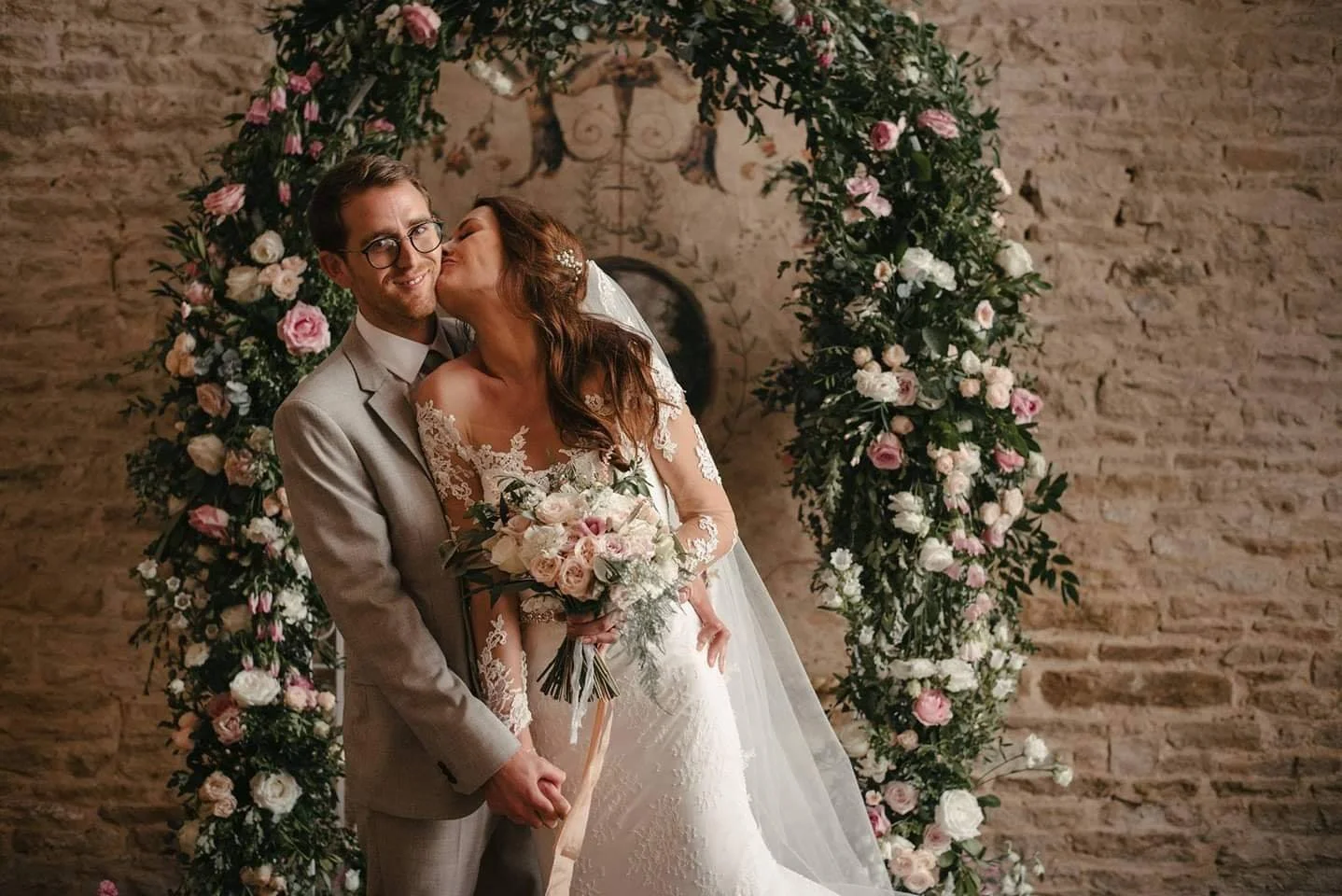 A bride and groom stand together in front of a floral wedding arch, with the bride kissing the groom on the cheek. The bride holds a bouquet of pink and white roses, and the groom wears a light grey suit with glasses. The background features a brick 