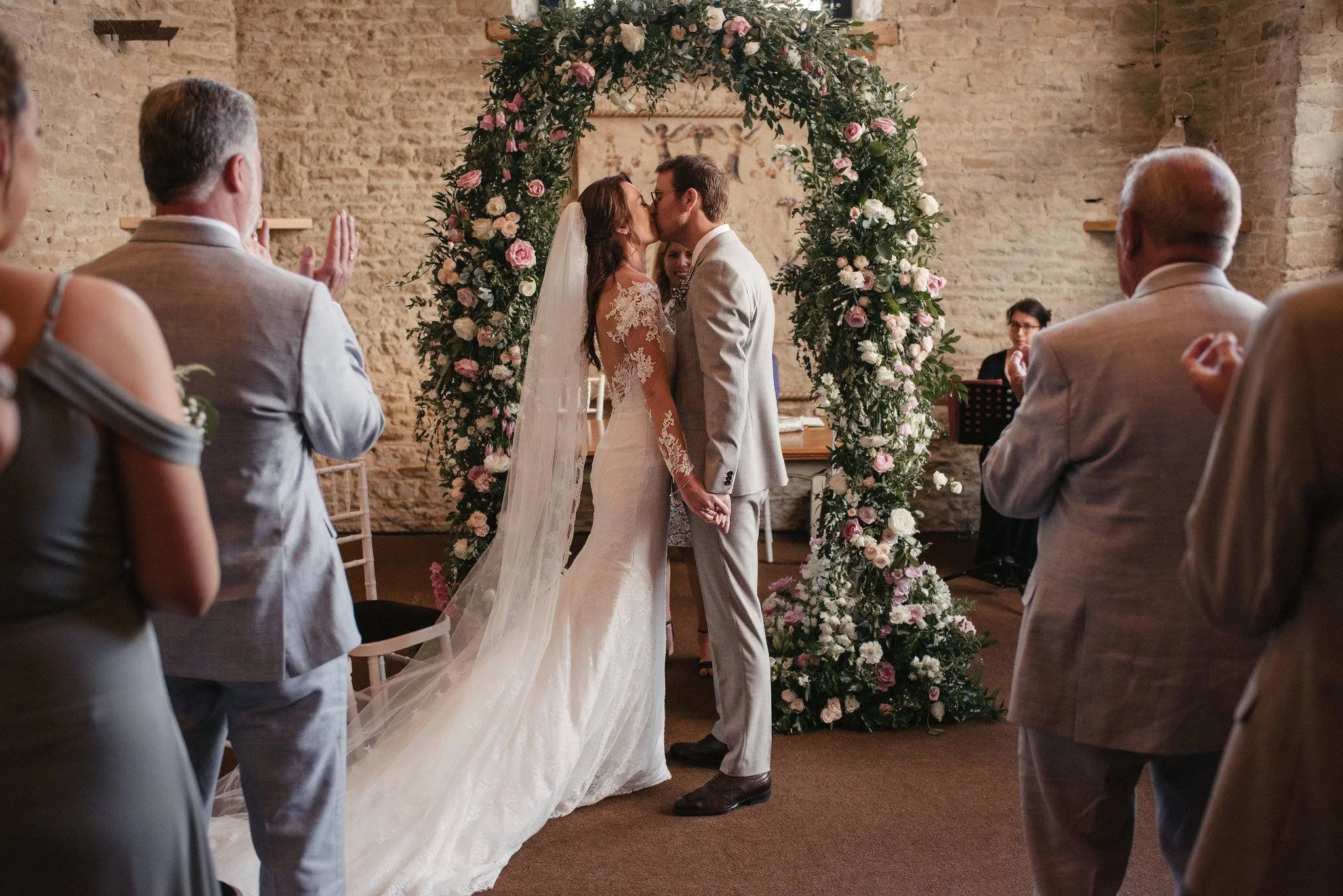 A couple kissing during a wedding ceremony in front of a floral arch, with guests watching and clapping.