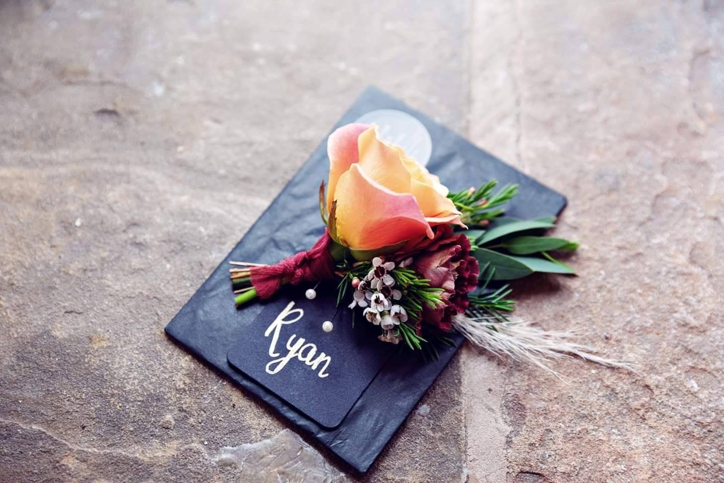 A boutonniere flower arrangement with a light pink rose, dark red flowers, greenery, white small flowers, and decorative feathers, placed on a black slate base with the name "Ryan" written on it.
