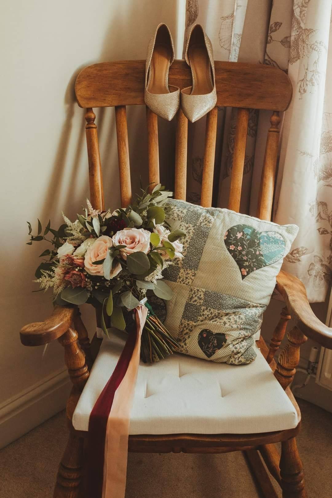 A pair of gold high-heeled shoes hanging on the back of a wooden chair, with a bouquet of flowers and a quilted pillow with heart and animal patterns placed on the seat.