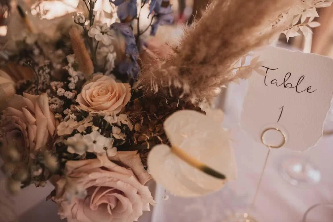 A floral centerpiece with blush pink roses, white flowers, dried foliage, and pampas grass, with a table number sign that reads 'Table 1' in elegant cursive.