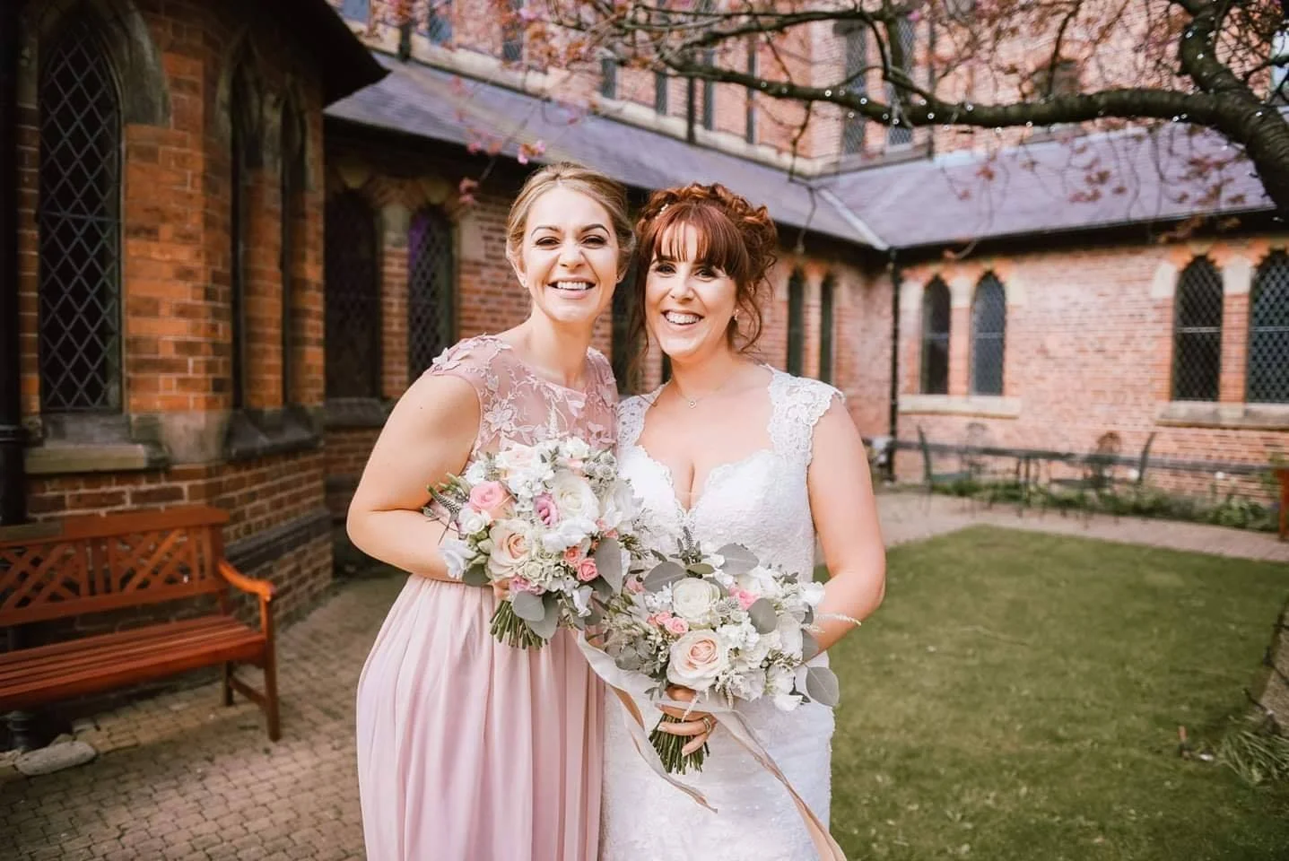 Two women smiling, one in a wedding dress holding a bouquet and the other in a bridesmaid dress holding a bouquet, standing outdoors in front of a brick building and trees with pink blossoms.