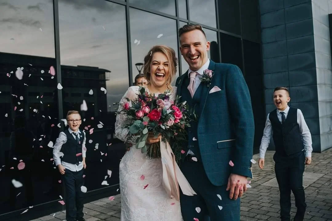 Happy bride and groom celebrating outside a modern building with children throwing flower petals in the air.