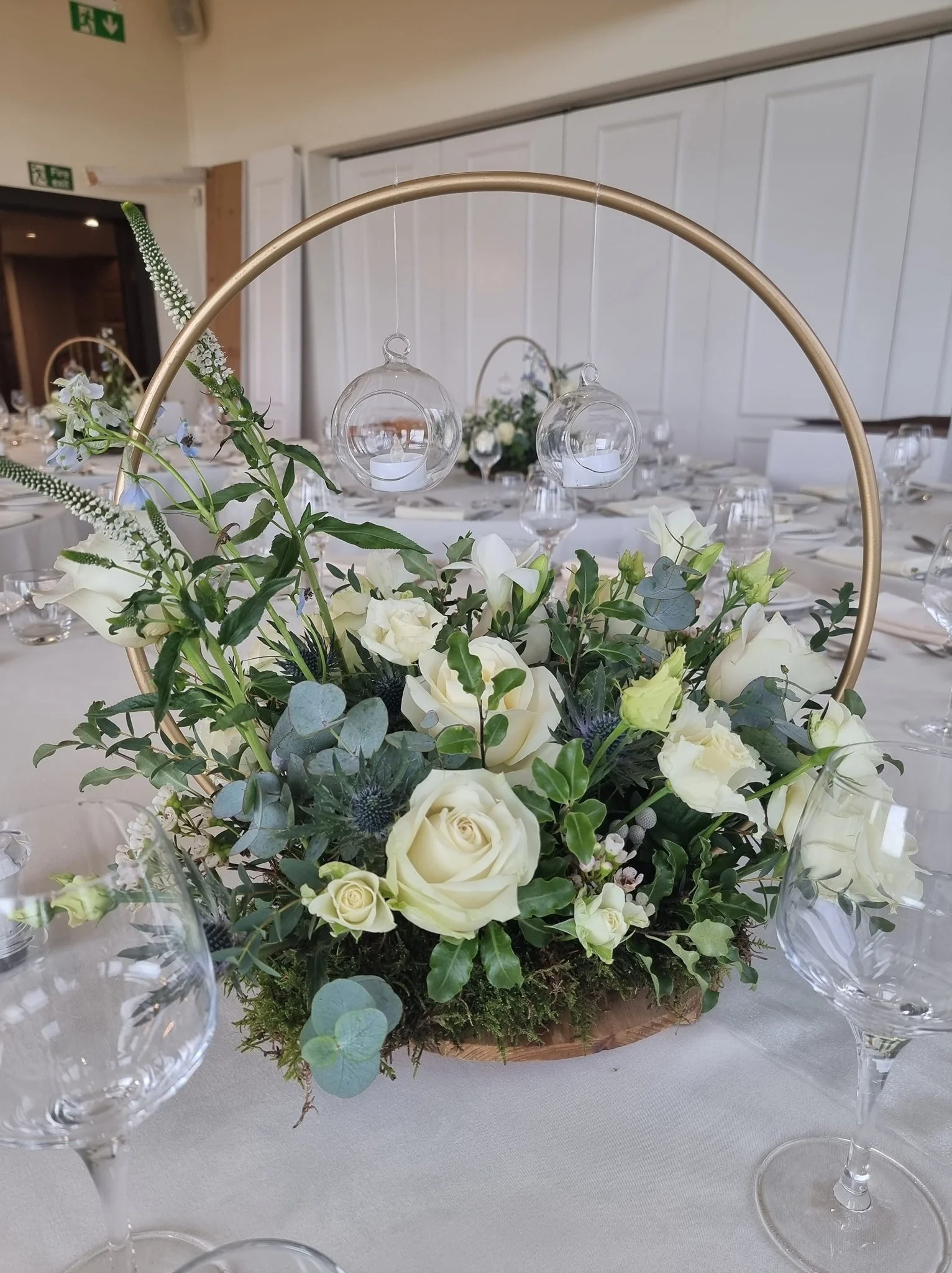 Elegant table centerpiece with white roses, greenery, and hanging glass candle holders at a wedding or event reception.