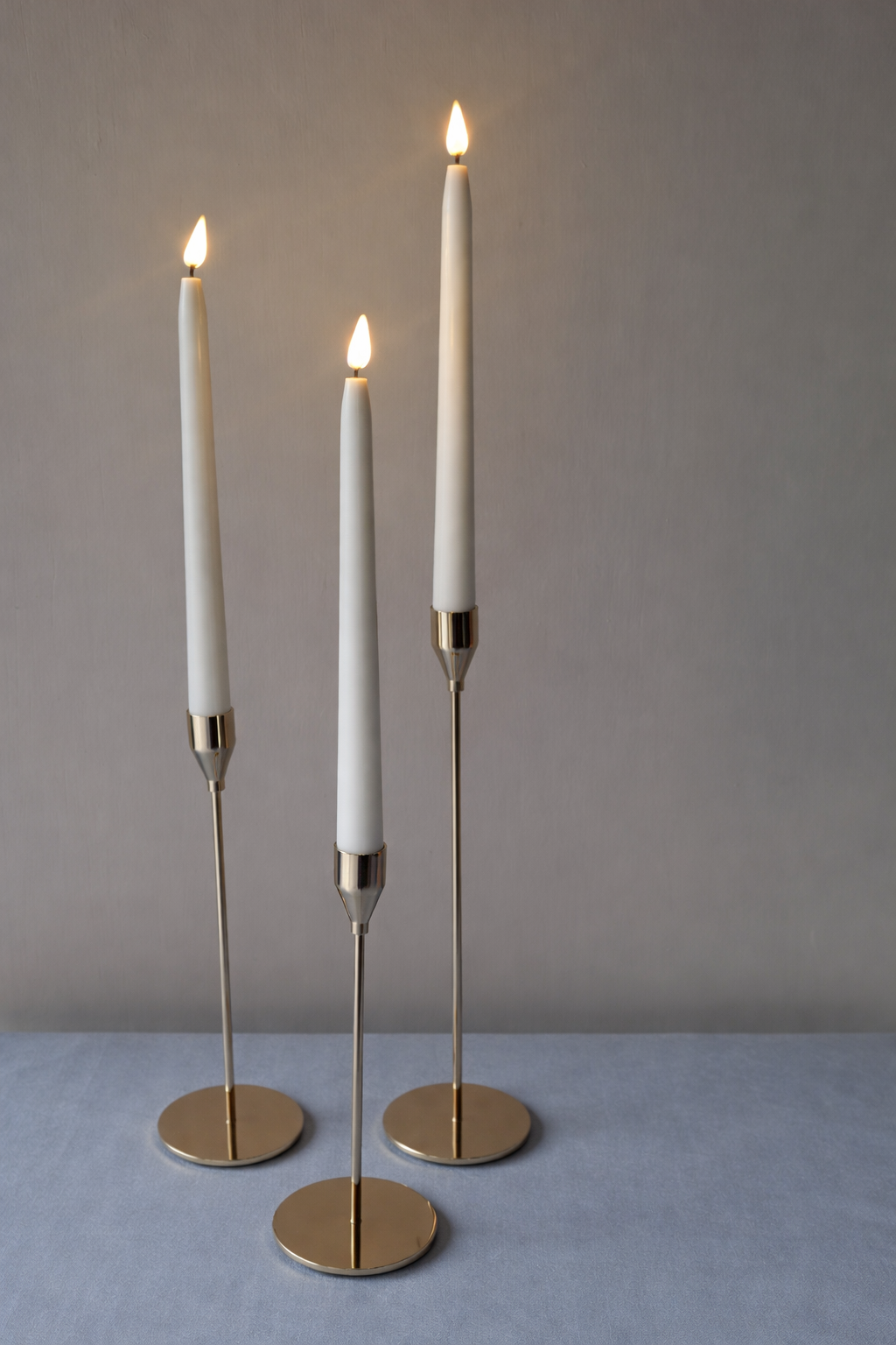Three modern white candlestick lamps with gold bases on a light blue surface, all with lit flames, against a neutral gray wall background.