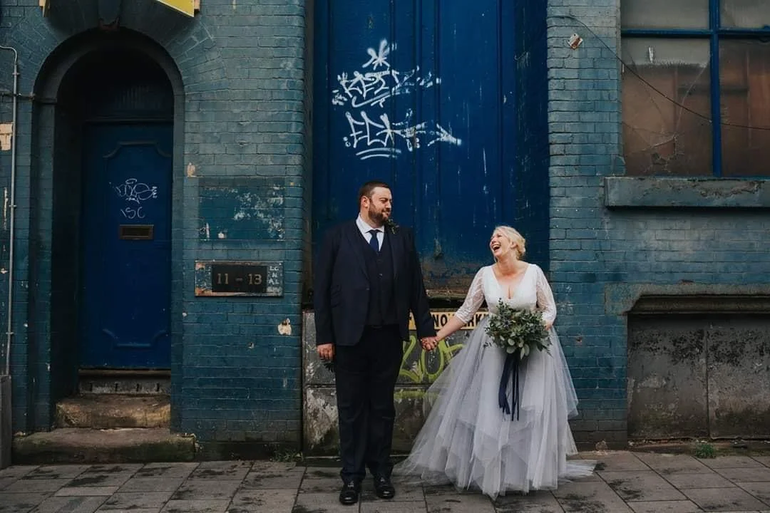 A newlywed couple holding hands and smiling in front of a blue graffiti-covered brick wall. The groom wears a dark suit and tie, and the bride wears a white wedding dress with a veil, holding a bouquet of flowers.
