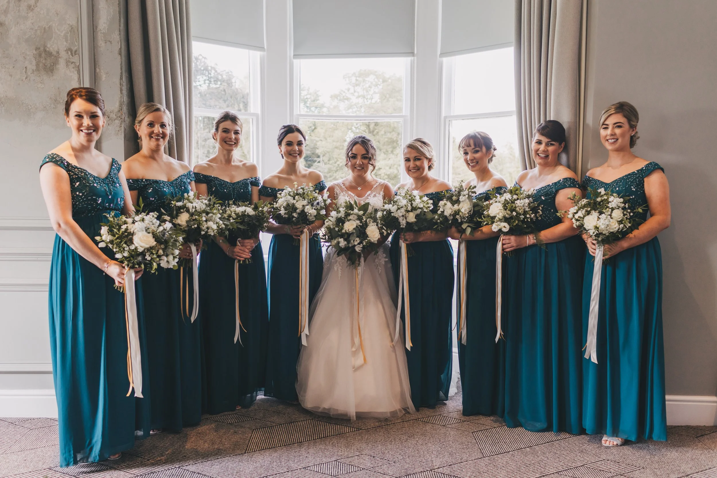 Bride and bridesmaids posing in front of large windows, holding bouquets of white flowers.