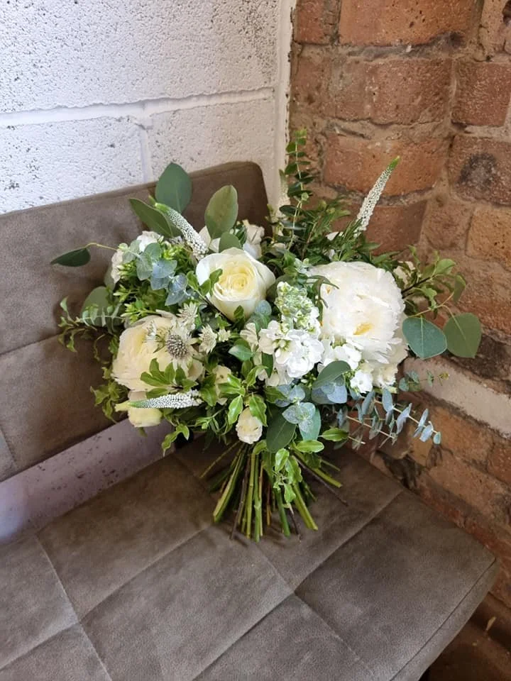 A bouquet of white roses, peonies, and various green foliage resting on a gray fabric bench next to a white painted brick wall and a red brick wall.
