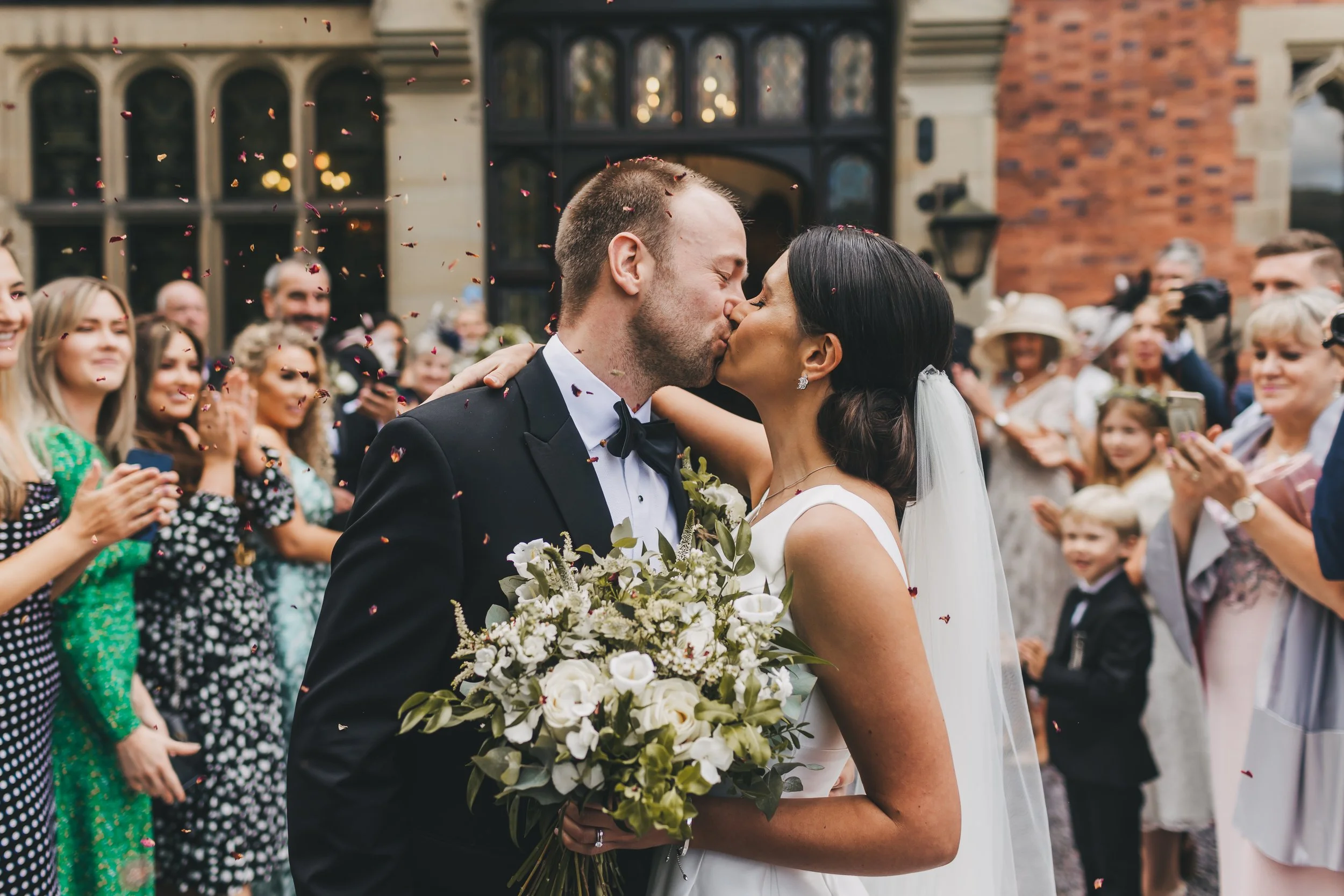 A bride and groom kiss at their wedding, holding a bouquet of white flowers amidst a crowd of guests celebrating.