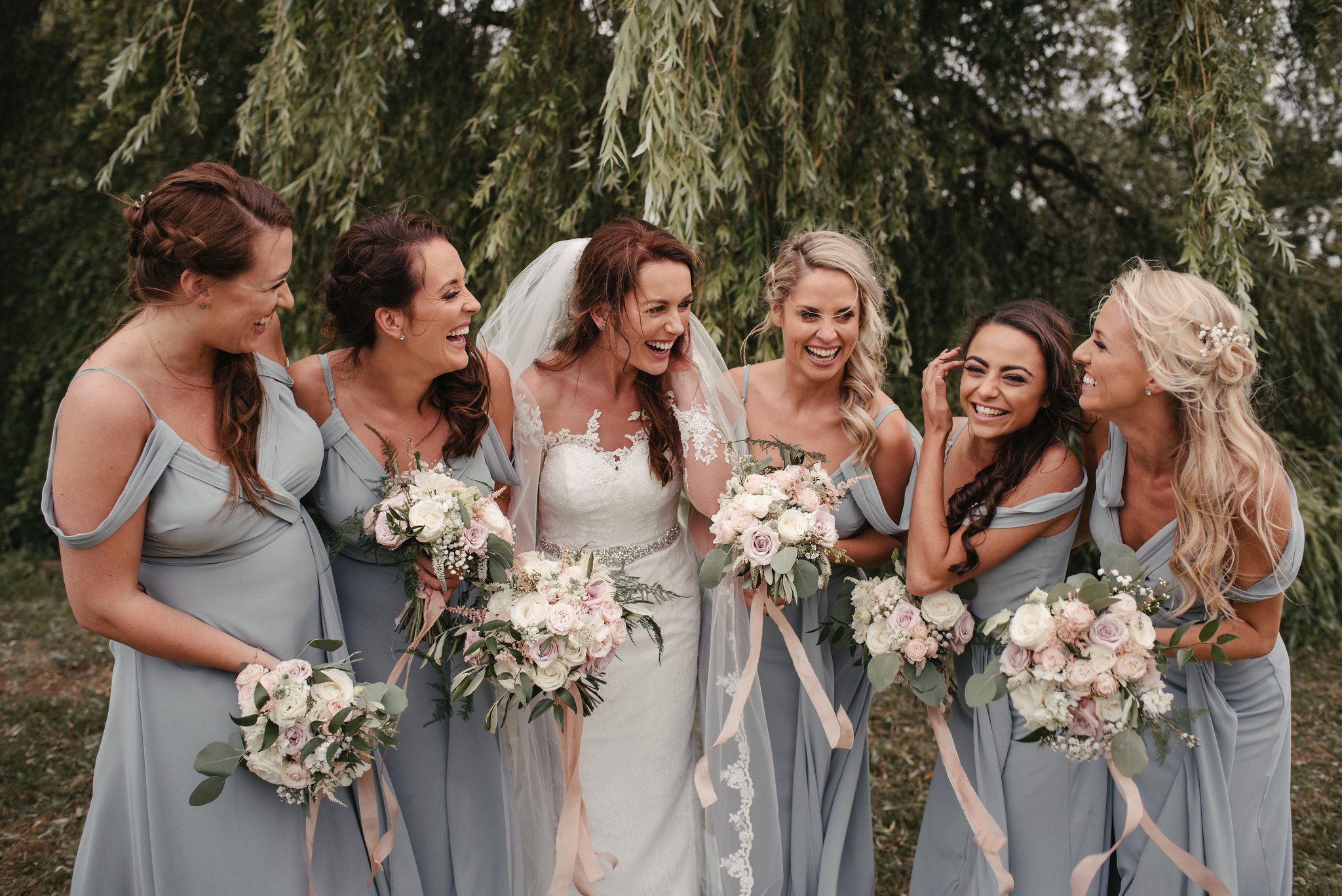 Bride and bridesmaids laughing together outdoors, holding bouquets of pink and white flowers, under a large leafy tree.