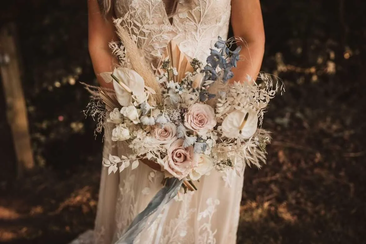 A woman in a lace dress holding a large bouquet of dried and fresh flowers, including roses, anthuriums, and eucalyptus, against a dark outdoor background.