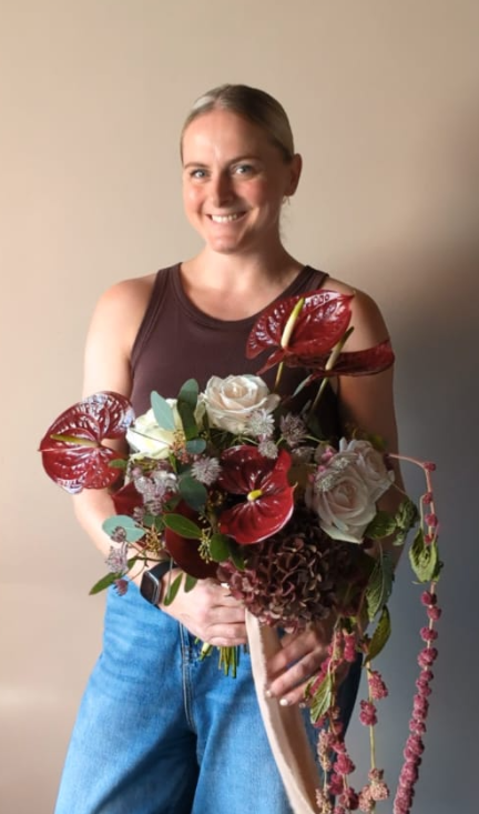 Woman holding a bouquet of flowers, smiling, wearing a sleeveless dark top and blue jeans, standing against a neutral background.