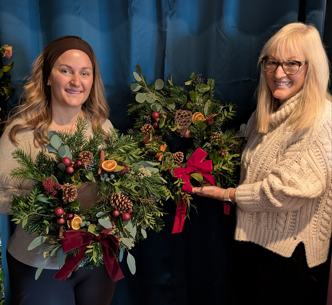 Two women holding holiday wreaths decorated with pinecones, berries, orange slices, and ribbons, standing in front of a blue curtain.