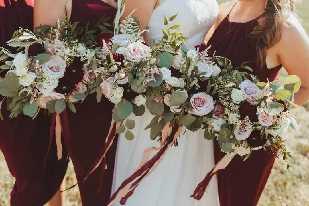 Close-up of a bride holding a large bouquet of blush pink, white, and deep red flowers with greenery, flanked by bridesmaids in burgundy dresses.