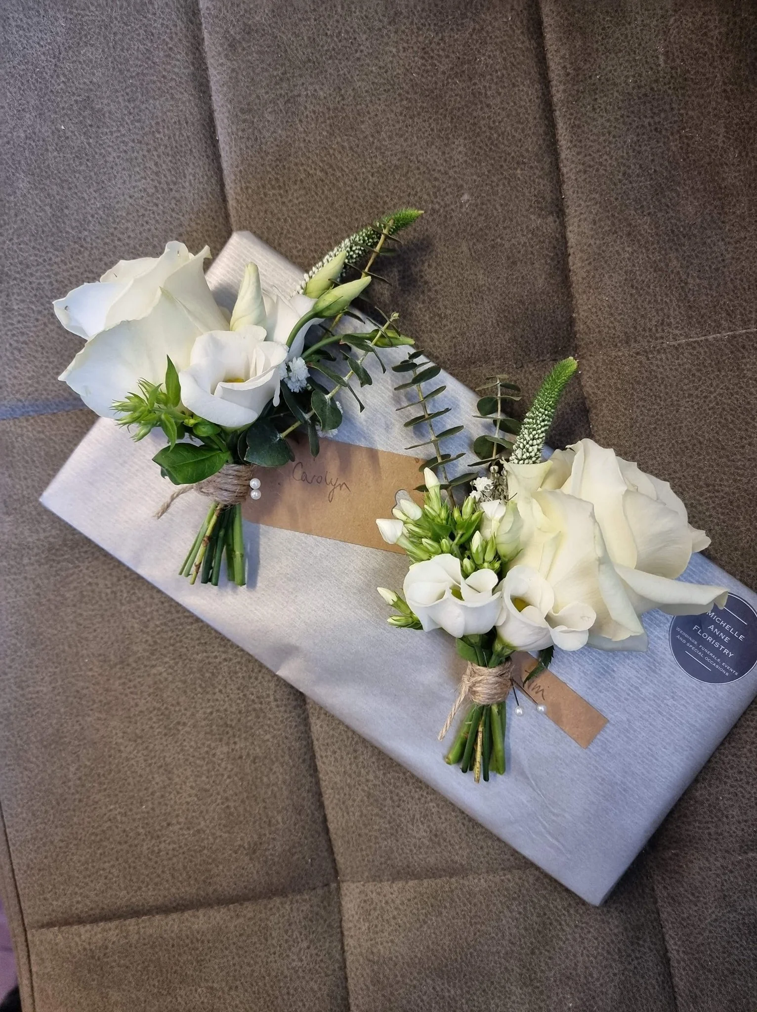 A floral arrangement consisting of white roses and lisianthus with greenery, attached to a gift wrapped in light-colored paper, placed on a brown textured surface.