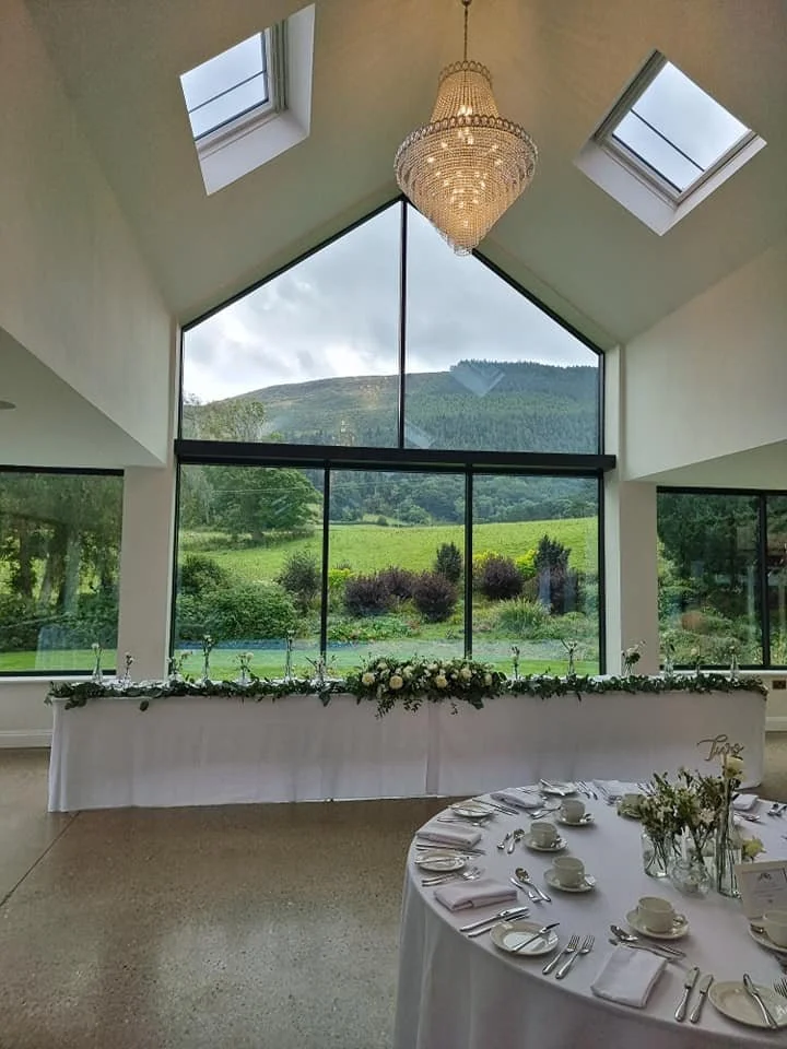 Interior of a reception hall with large glass windows and skylights, overlooking green hills and trees outside. A chandelier hangs from the ceiling. There is a long table decorated with flowers and greenery, and a round table set for a meal with whit