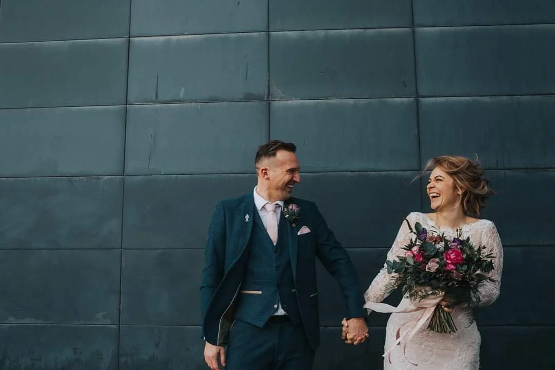 Happy wedding couple holding hands and smiling in front of a dark wall, woman in white dress holding a bouquet of flowers.