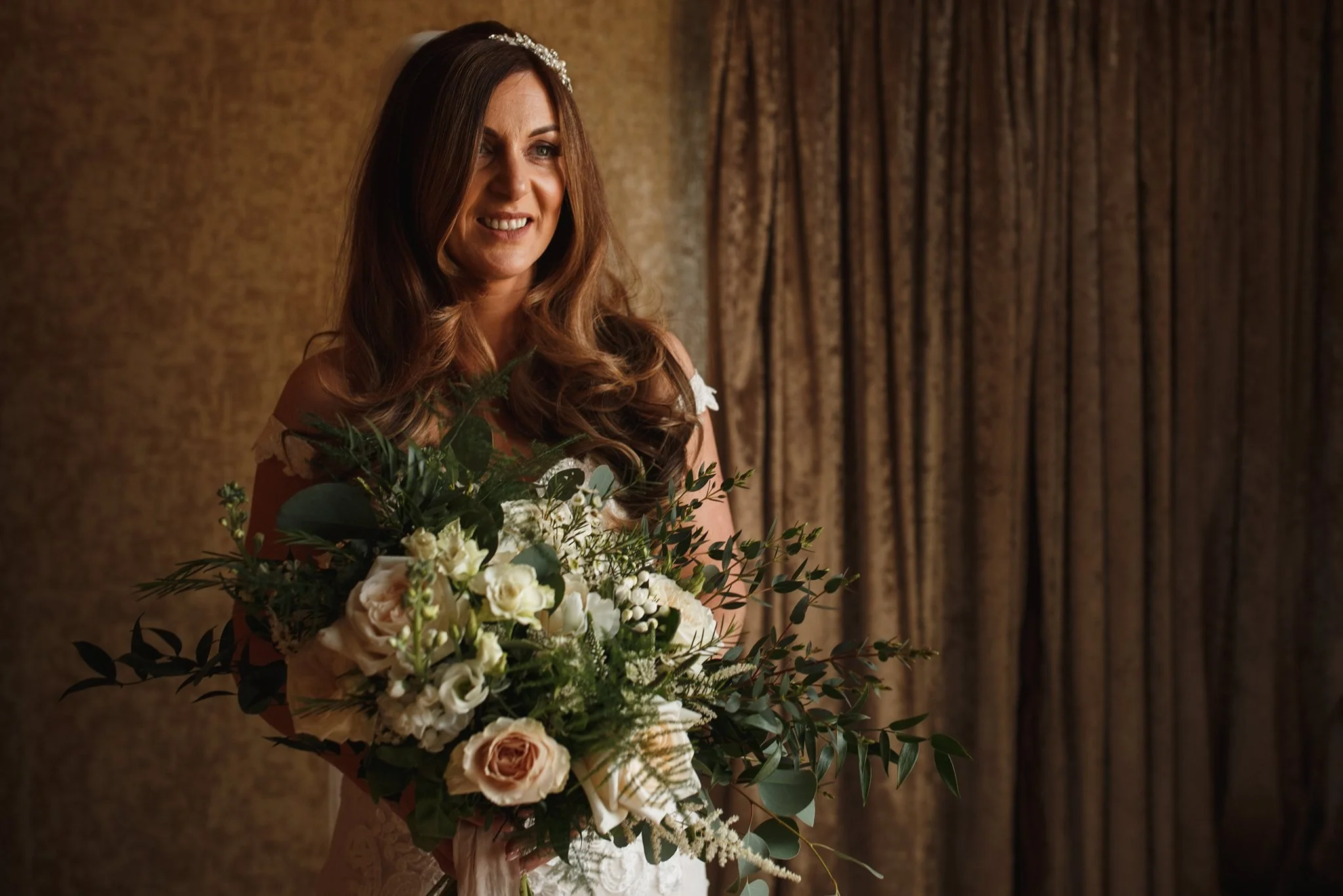 A smiling woman in a wedding dress holding a large bouquet of flowers, standing in front of a brown curtain.