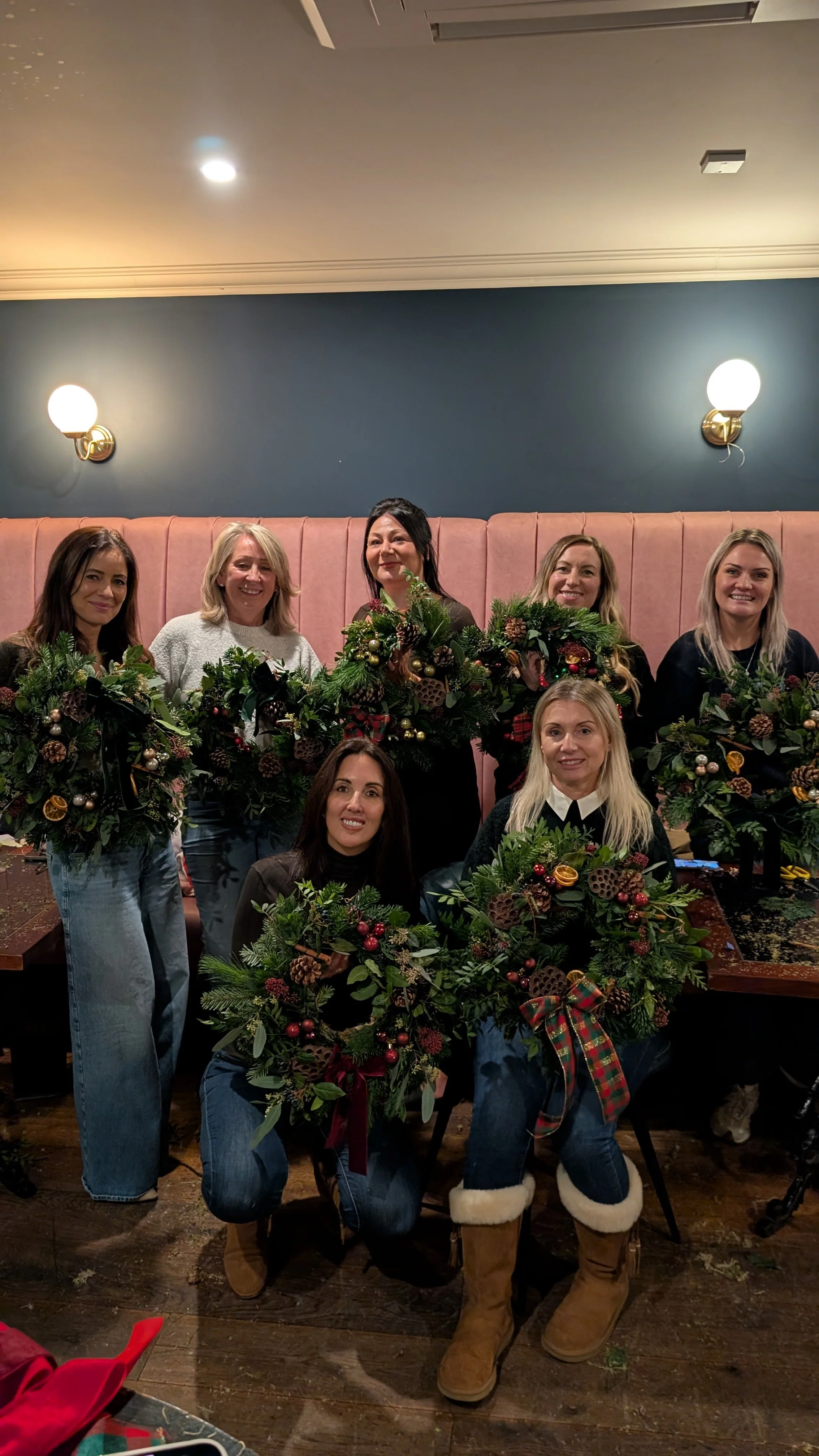 Group of women holding Christmas wreaths, gathered in a restaurant or cafe for a holiday celebration.