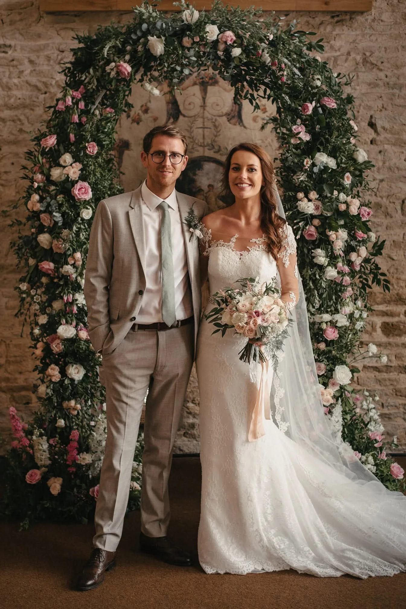A bride and groom posing for a wedding photo in front of a floral arch. The bride is holding a bouquet, wearing a lace wedding gown with a long veil, and smiling. The groom is dressed in a light suit with a tie and glasses, also smiling. The backgrou