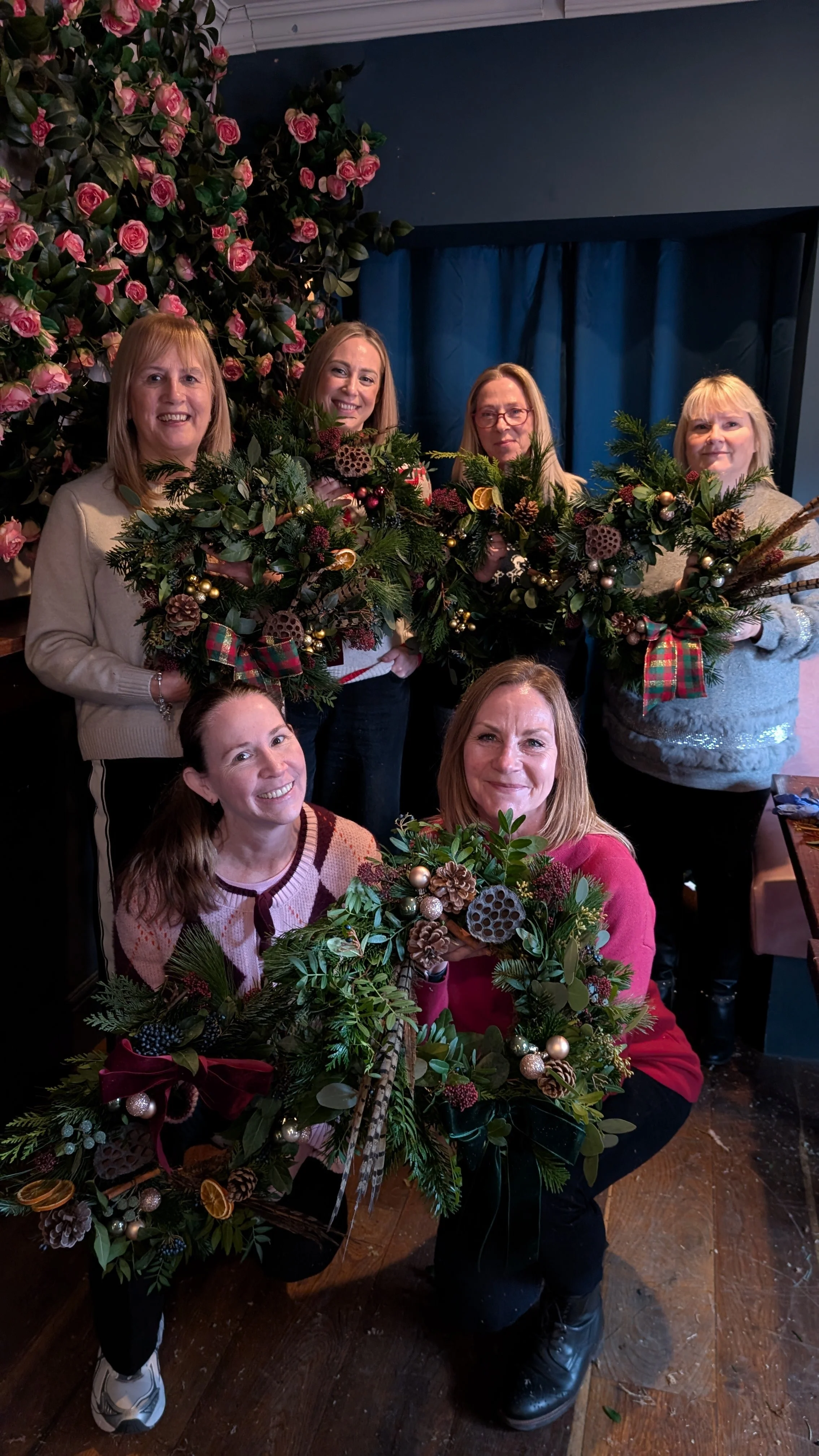 Group of six women holding festive Christmas wreaths decorated with pine cones, ornaments, and ribbons, standing indoors near a backdrop of pink roses and blue curtains.