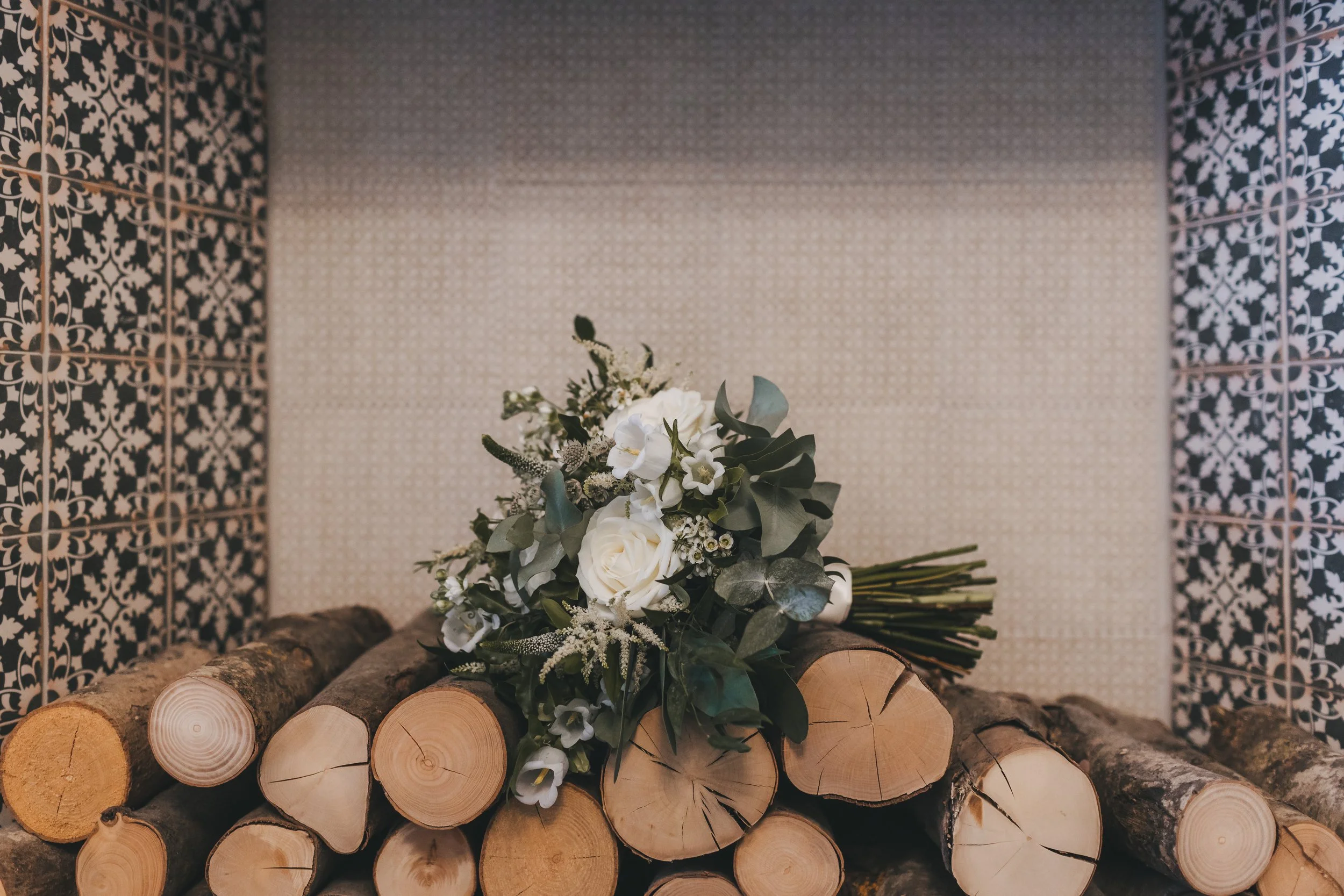 A bouquet of white flowers and greenery rests on a stack of cut wooden logs, with patterned wall tiles in the background.