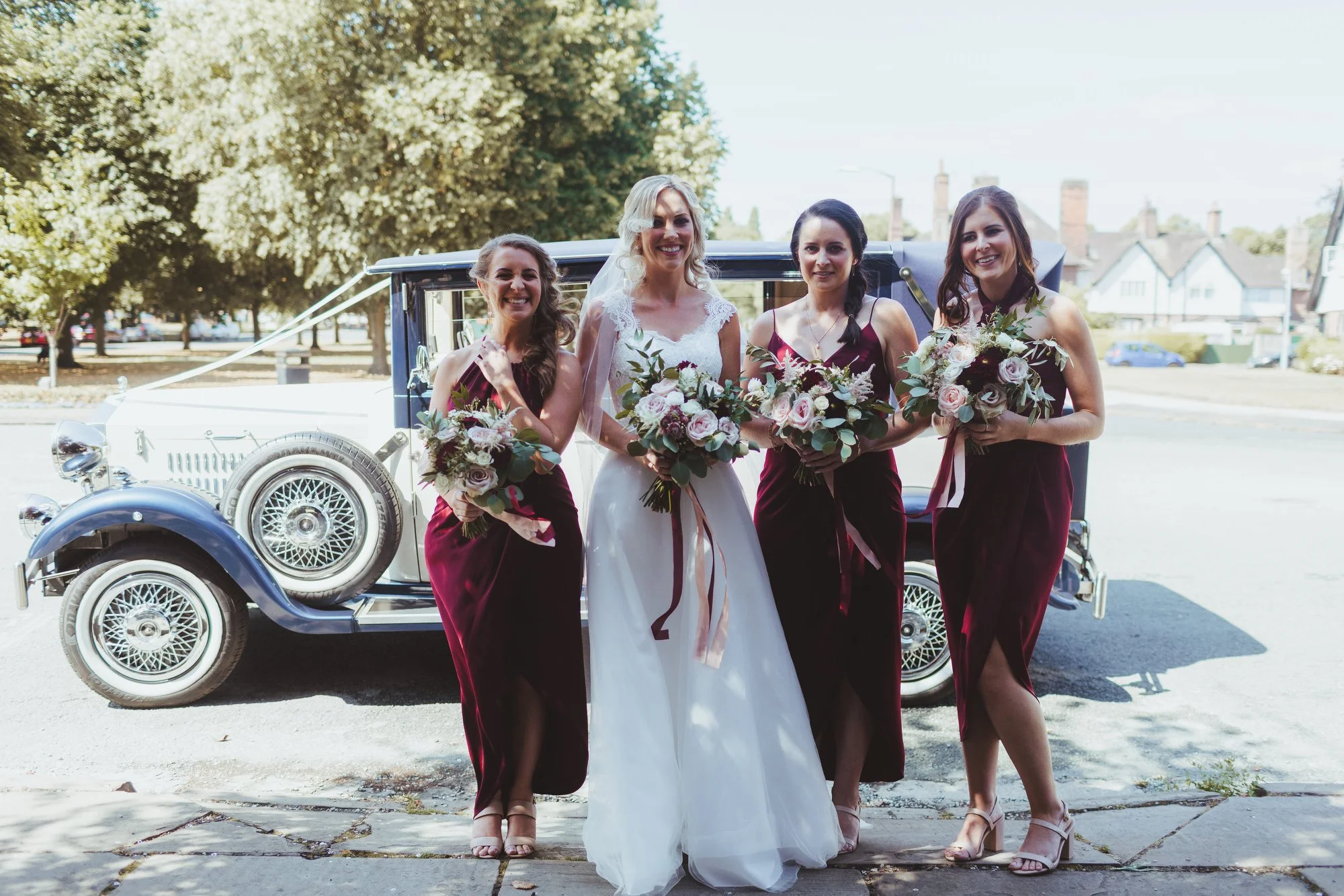 A bride in a white wedding dress and three bridesmaids in burgundy dresses standing in front of a vintage car, all holding bouquets of flowers.