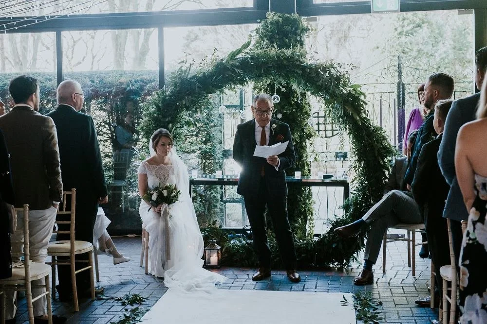 Wedding ceremony with a bride sitting in front of an officiant, surrounded by guests, under a floral arch in a glass-walled venue.